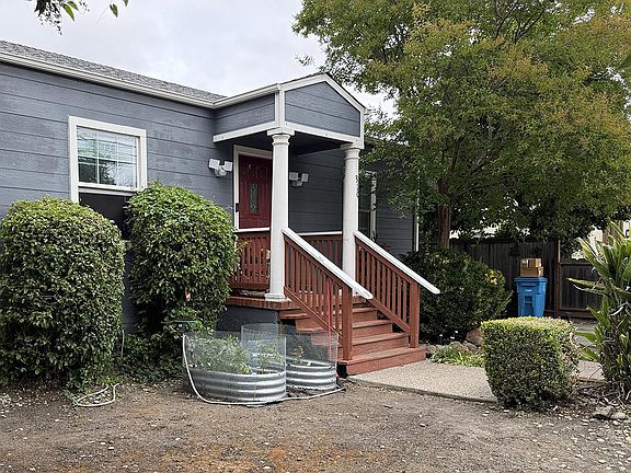 The gabled front porch of 310 Oak Ave, painted white with two columns framing a deep red front door, blue clapboard siding, and wooden steps down to the path