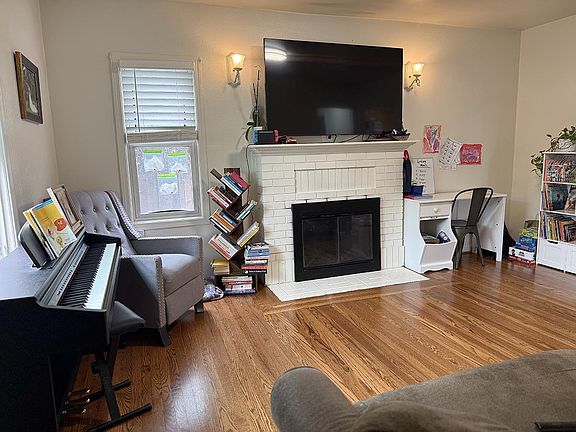 The living room at 310 Oak Ave with a white-painted brick fireplace, hardwood floors, an upholstered armchair, a piano along the left wall, and bookshelves