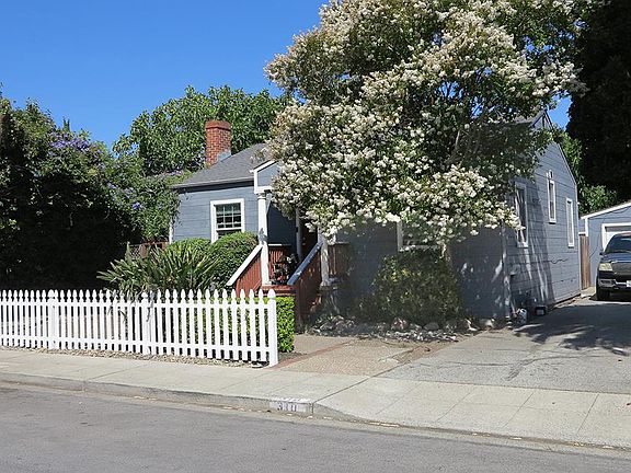 Front elevation of 310 Oak Ave, a 1937 blue clapboard cottage in Redwood City, with a brick chimney, white picket fence, and a crepe myrtle in full bloom over the front steps